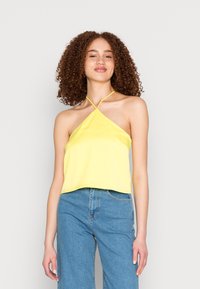 Top halter amarelo em cetim com um design de tiras cruzadas, apresentando um comprimento ligeiramente curto e uma textura suave, combinado com jeans azul de denim.