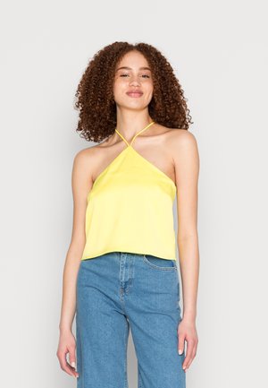 Top halter amarelo em cetim com um design de tiras cruzadas, apresentando um comprimento ligeiramente curto e uma textura suave, combinado com jeans azul de denim.