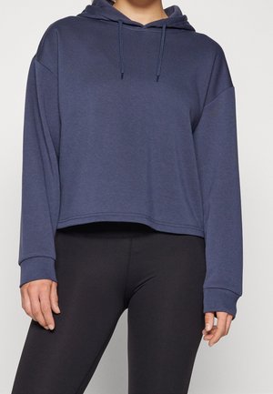 Person iført en marineblå cropped hættetrøje med snore og sorte tætsiddende leggings, stående mod en ensfarvet baggrund.
