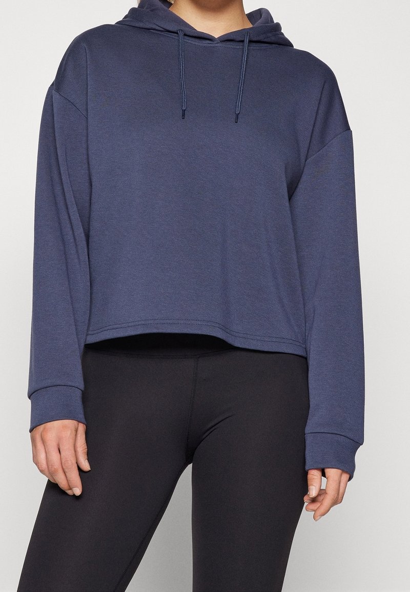 Personne portant un sweat à capuche court bleu marine avec cordons et des leggings noirs ajustés, debout devant un fond uni.