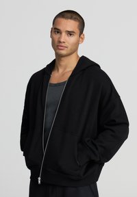 Felpa nera con zip realizzata in misto cotone, caratterizzata da un tessuto spesso, tasche frontali ampie e cappuccio con cordino. Indossata sopra una canotta grigia.