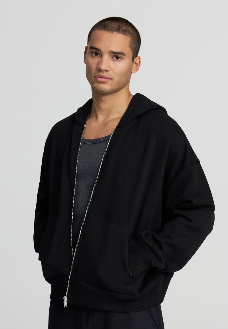 Felpa nera con zip realizzata in misto cotone, caratterizzata da un tessuto spesso, tasche frontali ampie e cappuccio con cordino. Indossata sopra una canotta grigia.