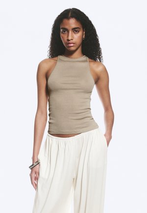 Woman with curly hair wearing sleeveless beige top and loose white pants, standing with one hand in pocket against plain background.