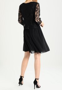 Kvinna klädd i svart spets långärmad topp och knä-lång svart kjol, stående med ryggen vänd, iförd svarta högklackade ankelskor med rem.