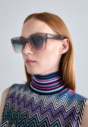 Yli suuret gradientti aurinkolasit, joissa on tumma kehys ja sävytetyt linssit, käytetään kuvioidun, monivärisen hihattoman paidan kanssa, jossa on korkea kaulus.