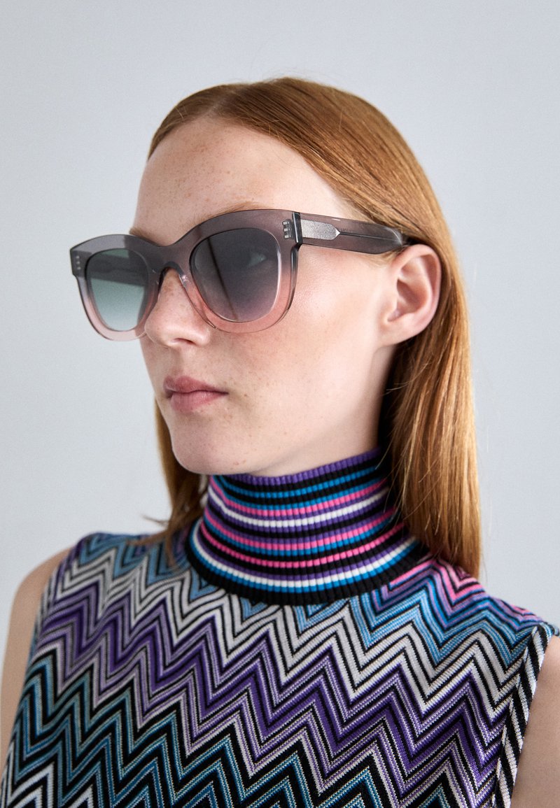Lunettes de soleil oversize dégradées avec une monture noire et des verres teintés, portées avec un haut sans manches à motifs multicolores comportant un col haut.