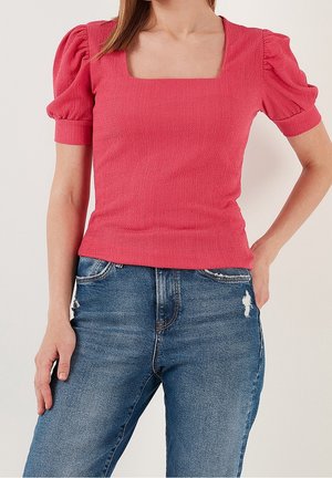 Kobieta ubrana w teksturowaną, jaskraworóżową bluzkę z bufkami i kwadratowym dekoltem oraz niebieskie, przetarte dżinsy, stojąca z jedną ręką w kieszeni.