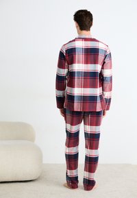 Hombre con pijamas de manga larga de cuadros en rojo, azul marino y blanco, de pie descalzo sobre una alfombra beige junto a un sofá curvo de color crema, de espaldas.