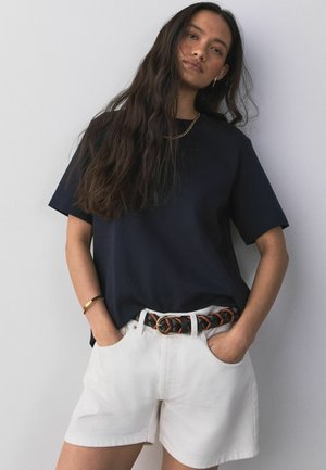 Pikajuustega naine, kes kannab musta lühikeste varrukatega särki, valgeid lühikesi pükse ja kootud musta-pruuni vööd, seisab käed taskutes.