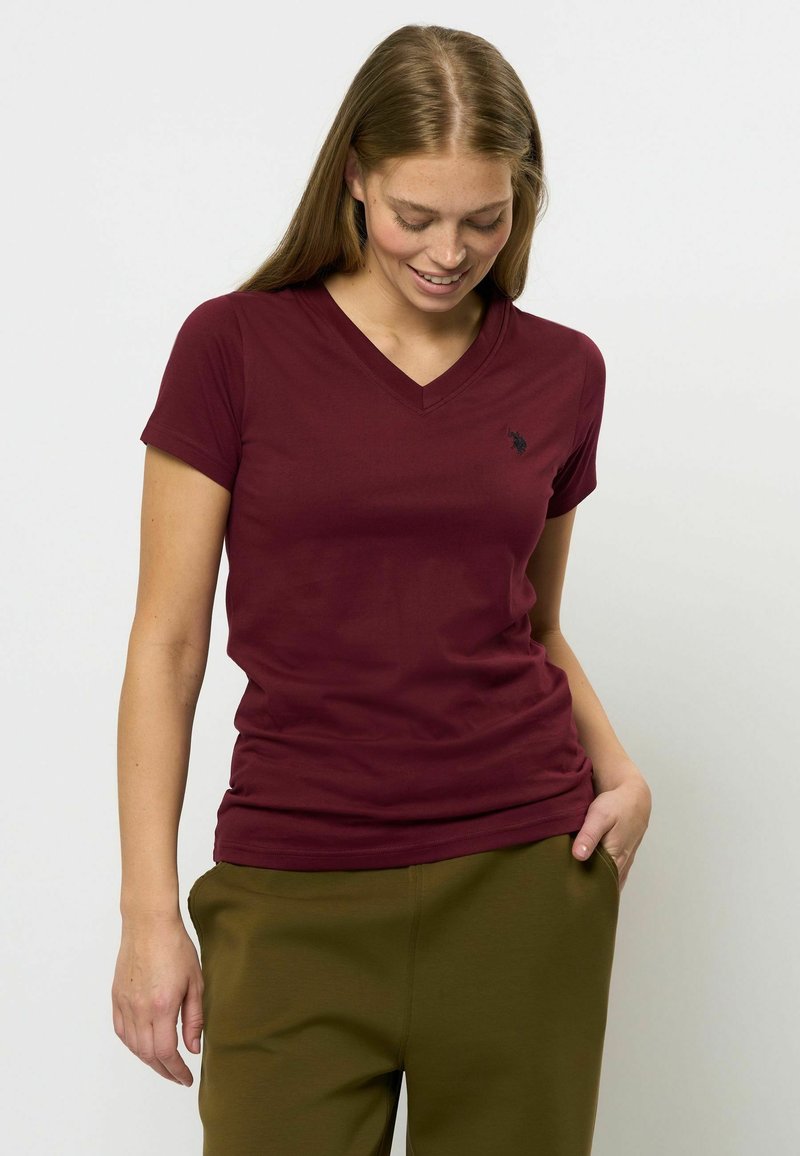 Burgundska v-neck majica iz mehkega materiala, s kratkimi rokavi in majhnim črnim logotipom na prsih. Nosena z olivno zelenimi hlačami.