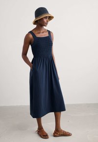 Navy blue sleeveless dress with smocked bodice and pockets, paired with a straw bucket hat and brown sandals on a neutral background.
