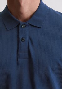 Marineblauwe polo met een gestructureerde stof, een knoopsluiting met twee knopen en een klassieke kraag. De knopen zijn donker en hebben een matte afwerking.