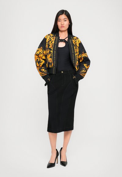 Veste bomber noire avec des motifs floraux dorés, assortie d'une jupe crayon noire avec des coutures visibles et de chaussures à talons hauts noires.