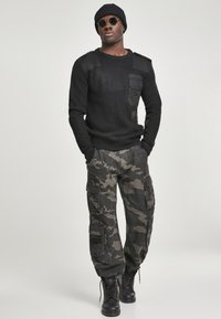 Suéter negro de punto con diseño texturizado y acentos en los hombros, combinado con pantalones cargo de camuflaje que cuentan con múltiples bolsillos. Botas negras de combate completan el conjunto.