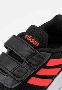 Černá atletická bota s prodyšným síťovaným svrškem, dvěma suchými zipy, červenými akcenty na značce a třemi pruhy. Bílá podrážka.