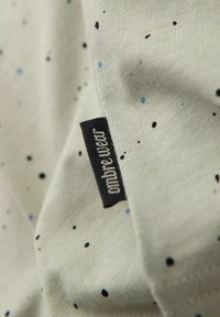 Tejido gris claro con pequeñas motas azules y negras. Presenta una etiqueta de tela negra con el texto "ombre wieter". Textura suave.