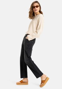 Suéter beige texturizado, jeans negros de cintura alta y sandalias de plataforma marrones. Gafas de sol marrones, manos en los bolsillos, perfil lateral.