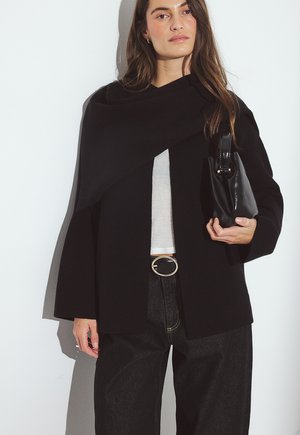 Donna che indossa un cardigan nero aperto sul davanti, top bianco, pantaloni neri con fibbia dorata e tiene in mano una piccola borsa nera su sfondo bianco.