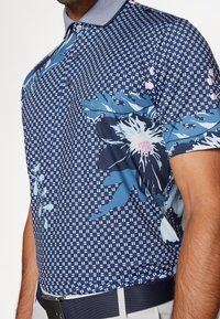 Polo de manga corta en tejido azul oscuro con un patrón geométrico y detalles florales en azul claro y rosa. Cuello con cierre de botones.