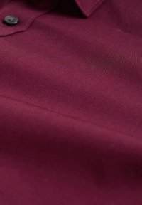 Burgundy tyg med en slät textur, utrustad med krage och en svart knapp, som visar fin sömnad längs kanterna.