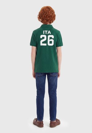 Παιδί με σγουρά κόκκινα μαλλιά που φοράει πράσινο πόλο με το «ITA 26» και μπλε τζιν, στέκεται με την πλάτη γυρισμένη σε απλό φόντο.