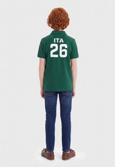 Παιδί με σγουρά κόκκινα μαλλιά που φοράει πράσινο πόλο με το «ITA 26» και μπλε τζιν, στέκεται με την πλάτη γυρισμένη σε απλό φόντο.