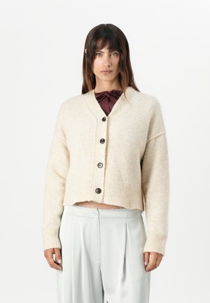 Gilet court de couleur crème en laine douce, avec un col en V, cinq boutons foncés et des poignets côtelés. Porté sur un pantalon gris pâle.