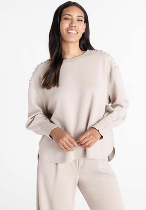 Vrouw glimlacht terwijl ze een beige top met lange mouwen draagt met parelaccenten op de schouders en bijpassende beige broek tegen een witte achtergrond.