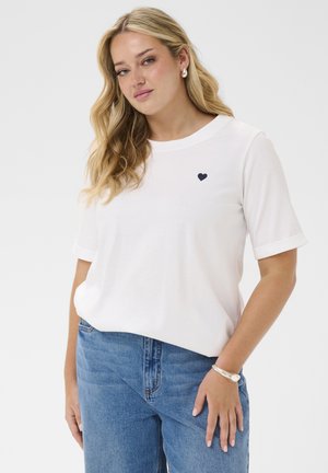 Jeune femme aux cheveux blonds portant un t-shirt blanc à manches courtes avec un petit emblème cœur noir et un jean bleu, posant devant un fond uni.