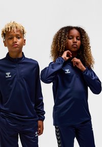 Marinblå sportpullover med halvlång dragkedja, tillverkad av slät tyg. Utmärkt logotyp på bröstet. Matchas med samordnade byxor med mönstrade accenter.