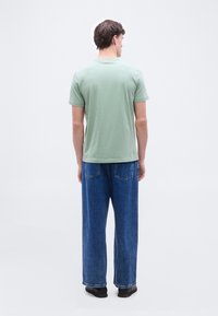 Άνδρας που στέκεται με την πλάτη, φοράει ανοιχτό πράσινο κοντομάνικο μπλουζάκι, φαρδιά μπλε τζιν και μαύρα παπούτσια, σε απλό λευκό φόντο.