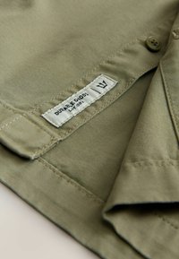 Olivgrünes Stoffhemd mit glatter Textur, versehen mit einem Etikett "DURABLE GOODS" und einem Knopfverschluss. Hochwertige Nähdetails.