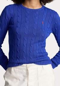 Blå kabelstickad tröja med rund halsringning och långa ärmar, med strukturerade mönster, avslutad med ribbad nederkant och liten logotypdetalj.