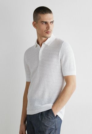 Mann trägt ein weißes kurzärmeliges, strukturiertes Strick-Poloshirt und blaue Hosen, steht mit einer Hand in der Tasche und schaut zur Seite.