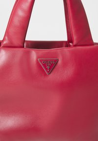 Borsa a tracolla in ecopelle rossa con doppi manici, patch triangolare in metallo con logo. Texture liscia, forma rettangolare compatta.