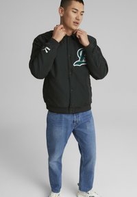 Svart bomberjakke med et grønt brodert "L"-logo, ribbede mansjetter og knappelukking, sammen med blå denimjeans og hvite joggesko.