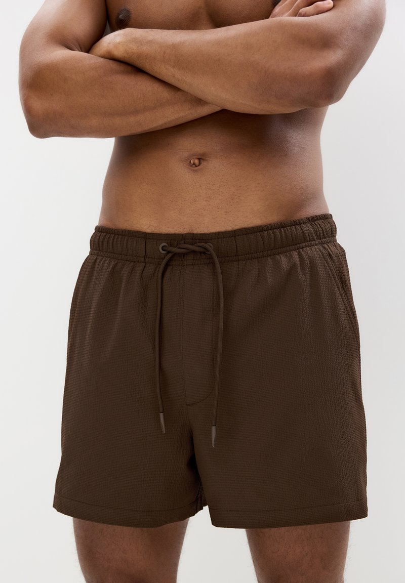 Braune Badeshorts mit elastischem Bund und Kordelzug. Texturiertes Material, knielange Ausführung, mit seitlichen Taschen und lockerer Passform.