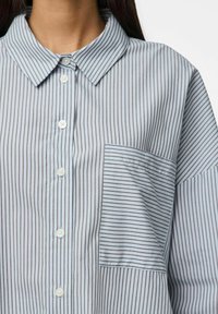 Košulja s gumbima u svijetloplavoj boji s bijelim uskim prugama, koja ima ovratnik, sedam gumba i lijevi džep na prsima s odgovarajućim prugama.