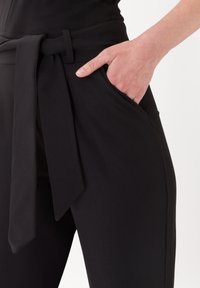 Černé kalhoty s širokým pasem a zavázaným opaskem, hlubokou boční kapsou a hladkým povrchem. Detailní pohled na ruku v kapse.