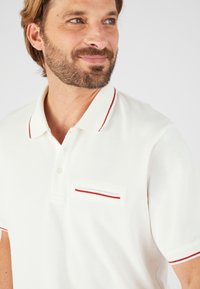 Camisa polo blanca con detalles rojos en el cuello y en el bolsillo. Presenta una tela texturizada y un único bolsillo en el pecho con un detalle de raya roja.