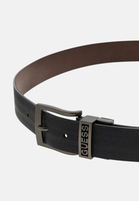 Sort gummibælte med fiskebensmønster, der har en metallisk spænde med "GUESS" branding. Glat inderside. Justerbart med huller.