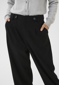 Black tailored trousers with front pleats, dual button closure, and side pockets; fabric appears smooth and structured. Paired with a gray cardigan.