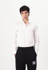 Witte button-up shirt met een gladde textuur, voorzien van een klein CK-logo op de linkerkant van de borst. Gecombineerd met zwarte sweatpants met elastische tailleband.