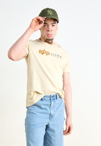 Bežs kokvilnas t-krekls ar "Alpha Industries" logo brūnā krāsā, apvienots ar gaiši ziliem džinsa šortiem. Zaļa beisbola cepure ar logo.