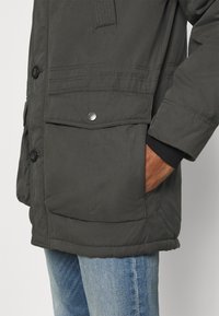 Personne portant une veste d'hiver vert foncé avec une grande poche avant à rabat et un jean bleu clair, la main glissée dans la poche de la veste.