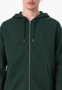 Sweat à capuche zippé vert en mélange de coton, avec une poche avant, des cordons de serrage et des accents en métal argenté.