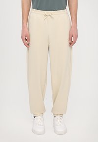 Beige sweatpants met een elastische tailleband en verstelbare trekkoord, met een relaxed fit. Gekleed met witte sneakers op een neutrale achtergrond.