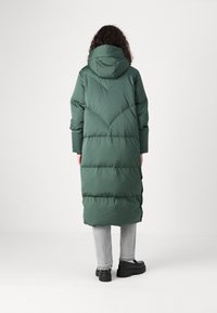 Grønn polstret jakke med hette, med en lang silhuett, quiltet design og synlige sømmer. Brukt over grå bukser og svarte sko.