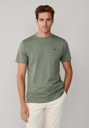 T-shirt vert à manches courtes en tissu doux avec un col rond et un petit logo noir sur la poitrine gauche. Porté avec un pantalon de couleur claire.