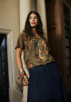 Woman with long dark hair leans against column wearing patterned brown shirt, navy skirt, layered necklaces, and floral scarf tied around wrist.